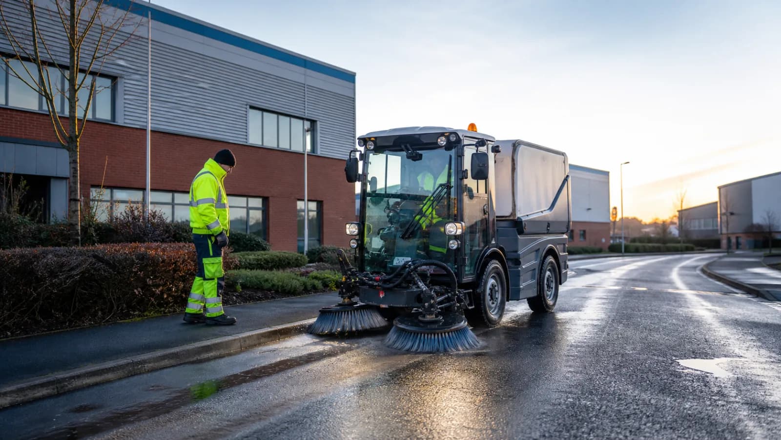 Compact road sweeper and operator cleaning a wet road on a UK industrial estate